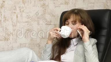 女商人坐在椅子上，光着脚在桌子上放松，喝茶，喝咖啡。 在办公室工作的女孩